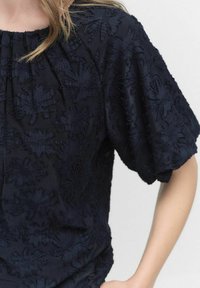 Marineblå broderet top med tekstureret blomsterdesign, korte puffærmer og rynket halsudskæring. Blødt stof med en afslappet pasform.