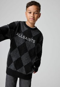 Ragazzo che indossa un maglione argyle nero e grigio con il testo "ALLSAINTS", in piedi contro uno sfondo grigio chiaro uniforme.
