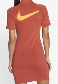 Sukienka o dopasowanym kroju z krótkim rękawem w kolorze rdzy, z wysokim kołnierzem i dużym żółtym logo Nike na plecach. Wykonana z gładkiego, elastycznego materiału.