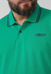 Groene polo met een textuurstof, voorzien van een knoopsluiting met twee knopen, korte mouwen en marineblauwe gestreepte accenten op de kraag. Merkenlogo aanwezig.