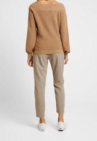 Beżowy prążkowany sweter z odsłoniętymi ramionami, zestawiony z jasnobrązowymi, kratkowanymi spodniami. Modelka ma na sobie białe buty, widziana od tyłu.