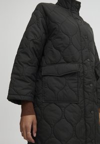 Abrigo negro acolchado con cuello alto, diseño sobredimensionado, gran bolsillo parche y cierre con botones a presión. Presenta un patrón texturizado y burbujeante.