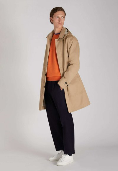 Beige Trenchcoat mit Kragen, zwei vorderen Taschen, über einem orangefarbenen Pullover und einer marineblauen Hose getragen; kombiniert mit weißen Sneakern.