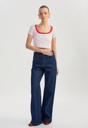 Lyserød ribbet crop top med rød kant, kombineret med blå vide denimjeans. Modellen bærer røde sko; minimal baggrund.