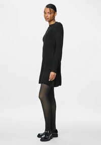 Abito nero a maglia con maniche lunghe, texture a costine, leggero dettaglio all'orlo inferiore, abbinato a collant neri e scarpe chunky ben lucidate.