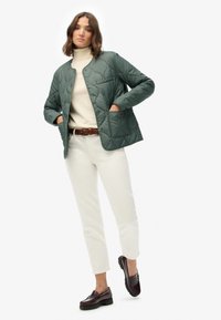 Groene gewatteerde jas met drukknoopjes, twee zakken, gedragen over een crèmekleurige coltrui en witte broek, gecombineerd met donkere instappers.