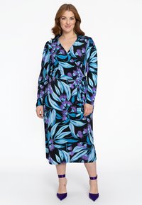 Schwarzes, langärmliges Wickelkleid mit blauem und lila Blumenmuster. Verfügt über einen Bindegürtel und einen knöchellangen Schnitt, kombiniert mit lila Absätzen.