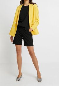 Yellow blazer with silver buttons, black top and shorts, gray and white patterned heels. Smooth fabric, tailored fit, modern design.