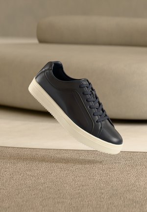 Zapatilla deportiva de cuero negro con suela y cordones blancos, flotando sobre una alfombra beige y un sofá de color claro al fondo.
