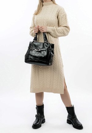 Femme en robe midi beige en maille torsadée, tenant un sac à main noir texturé, portant des bottes noires mi-mollet avec des boucles sur fond blanc.