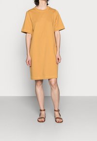Persona vestida con un vestido suelto de color mostaza a la altura de la rodilla y sandalias marrones con correa de tobillo, de pie contra un fondo claro y liso.