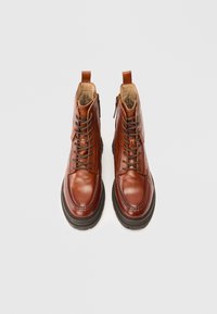 Brune skinnsnørte støvler med polstrede interiører og tykke sorte såler, sett ovenfra på en lys grå bakgrunn.