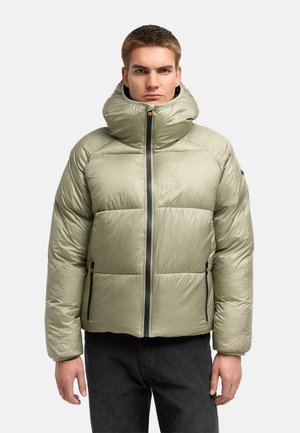 Giovane uomo che indossa una giacca invernale imbottita verde chiaro con la cerniera chiusa, in piedi davanti a uno sfondo chiaro e uniforme.