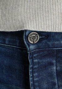 Vaqueros de mezclilla azul oscuro con un botón metálico visible que lleva la inscripción "PME LEGEND", y un suéter gris acanalado que cubre parcialmente la cintura.