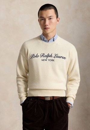 Homme portant un pull crème avec le texte "Polo Ralph Lauren New York", chemise bleue à col, pantalon foncé et ceinture marron, debout les mains dans les poches.