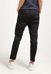 Czarne spodnie w dopasowanym kroju z przednimi kieszeniami. Materiał jest gładki, z delikatnym połyskiem. Noszone z białymi sneakersami, na których widać tatuaż nad kostką.