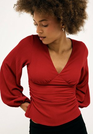 Vrouw met krullend haar die een gestructureerde rode blouse draagt met lange pofmouwen en een V-hals, kijkt naar beneden aan haar linkerzijde.