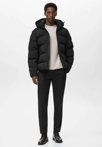 Giacca a vento nera con cappuccio, caratterizzata da una chiusura frontale con zip, texture imbottita e un design aderente. Indossata sopra una maglia grigia chiara e pantaloni scuri.