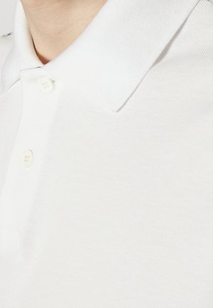 Polo blanc avec col en bord-côte, comportant une patte à deux boutons, tissu texturé et finition lisse.