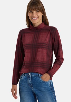 Femme souriante portant un col roulé à manches longues à carreaux bordeaux et un jean bleu, une main dans la poche, sur un fond uni.