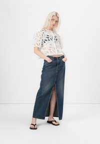 Haut court en dentelle florale blanche avec des manches bouffantes associé à une longue jupe en denim foncé avec une fente à l'avant. Le modèle porte des tongs noires.