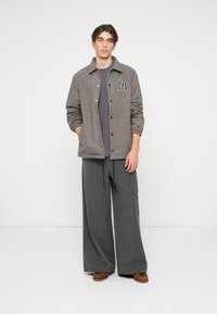 Jachetă în carouri maro și negre cu nasturi, tricou gri, pantaloni largi cu dungi subțiri gri și pantofi maro. Design minimal.