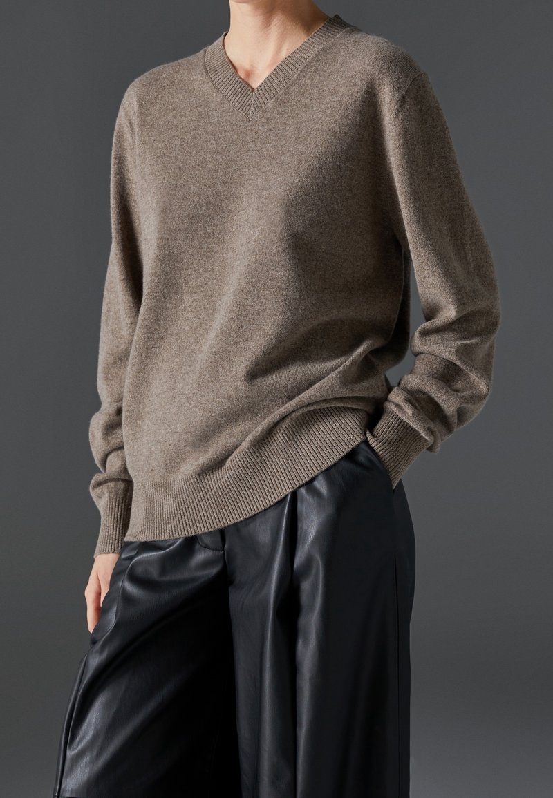 Personne portant un pull taupe à col en V et un pantalon large en cuir noir, debout avec une main dans la poche devant un fond gris foncé.