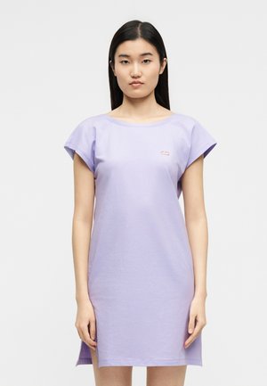 Pikkade tumedate juustega naine, kes kannab lühikeste varrukatega helelillat keskmise pikkusega kleiti, mille rinnal on väike logo, taust on lihtne.