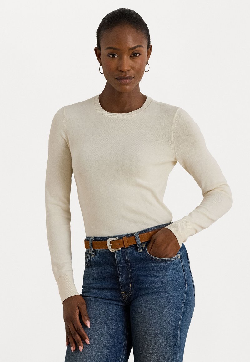 Maglione leggero beige a maniche lunghe con scollatura rotonda, design aderente e polsini a costine, abbinato a jeans blu scuro e una cintura marrone.