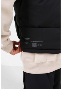 Schwarze Nylonweste mit gesteppten Abschnitten und einem Etikett mit QR-Code. Darunter getragen ein cremefarbener Pullover mit gerippten Bündchen und Saum.