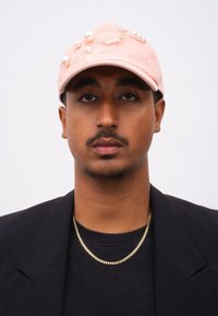 Rosa Baumwollmütze, verziert mit weißen Perlen und goldenen Sicherheitsnadeln, mit einem gebogenen Schirm und einer strukturierten Form. Schlichter Hintergrund.