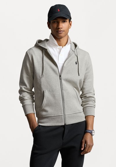 Grijze zip-up hoodie gemaakt van stof met een gladde textuur, voorzien van een buidelzak en een klein logo op de linkerborst. Zwarte pet gedragen.