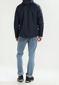 Mörkblå vattentät jacka med huva, har en slät yta och avslappnad passform, tillsammans med ljusblå jeans och svarta träningsskor.