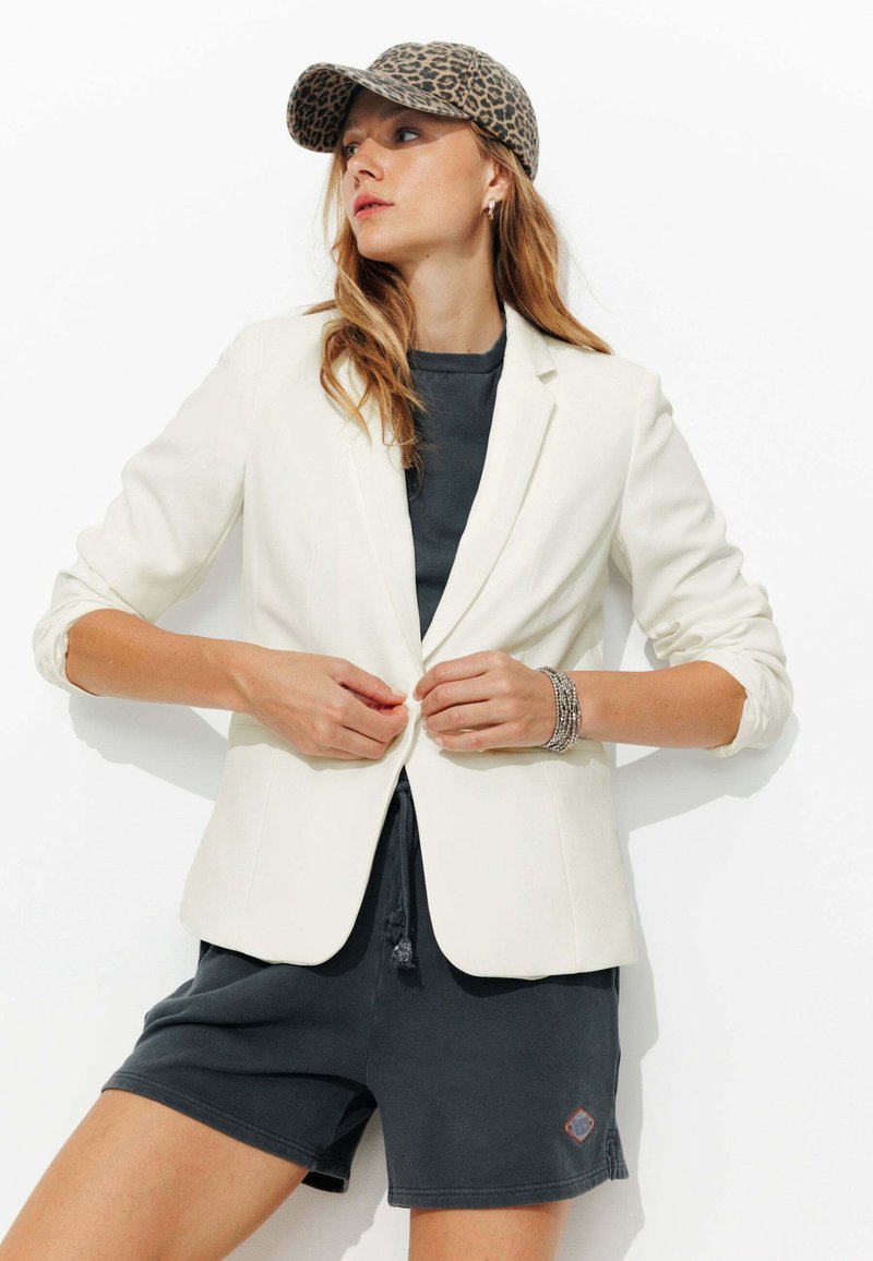 Blazer blanc sur mesure avec un bouton, porté sur une chemise gris foncé et un short. Accentué par une casquette en imprimé léopard. Texture de tissu lisse.