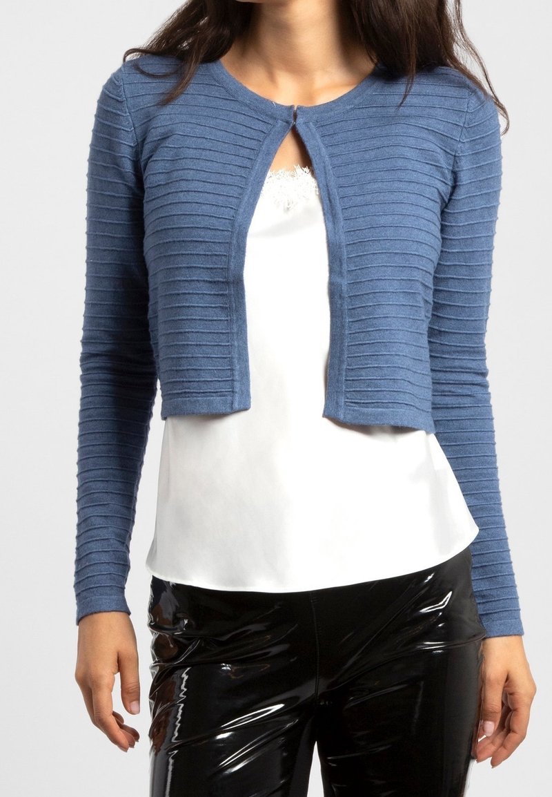Femme portant un cardigan court côtelé bleu sur un haut blanc bordé de dentelle et un pantalon noir brillant, debout devant un fond uni.