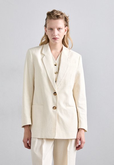 Texturierter cremefarbener Blazer mit einem Reverskragen, drei braunen Knöpfen und seitlichen Taschen, getragen über einem passenden Oberteil und lockeren Hosen.