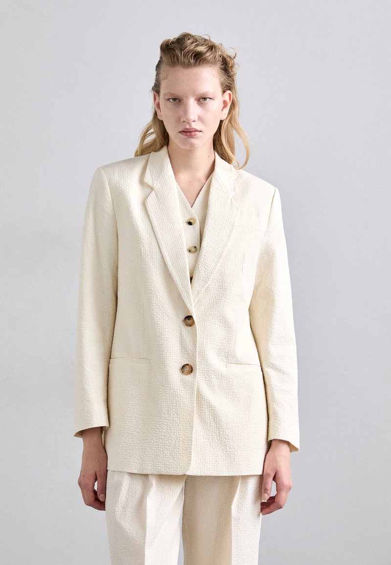 Geweven crème blazer met een gekraagde revers, drie bruine knopen en zijzakken, gedragen over een bijpassend topje en losse broeken.
