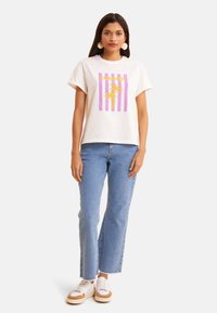 Camiseta de manga corta crema con gráfico en púrpura y amarillo de "VERANO" y diseño de palmera. Combinada con jeans de mezclilla claros y zapatillas.