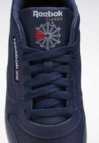 Tamsiai mėlyni odiniai sportbačiai su plokščiomis raištelėmis, perforuota pirštų dėžė, Reebok logotipas ir raudonos bei baltos akcentai ant liežuvėlio. Guminis padas.