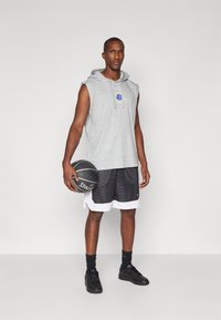 Pelēks bezpiedurkņu kapuce ar priekšējo logo, apvienots ar melniem sieta basketbola šortiem ar baltu apmalīti un melnām trenēšanās kurpēm, turētājs basketbola.