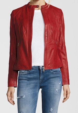 Vrouw die een nauwsluitend rood leren jack draagt over een wit shirt met verweerde blauwe jeans, staand met de armen ontspannen langs haar zij.