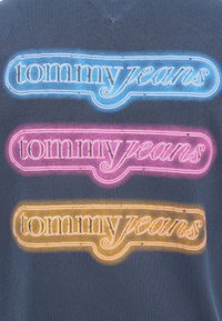 Σκούρο γκρι μπλουζάκι με τρία στοιβαγμένα λογότυπα "tommy jeans" σε μπλε, ροζ και πορτοκαλί neon στυλ με περίγραμμα.