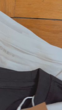 Pantalones en blanco y negro apilados sobre una superficie de madera; el negro tiene una cinturilla con una etiqueta adjunta, mientras que el blanco tiene una textura suave.
