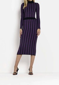 Langärmliges Midi-Kleid im lila und schwarzen Hahnentritt-Muster, mit taillierter Silhouette und schwarzem Taillenband. Getragen mit schwarzen High-Heel-Sandalen.