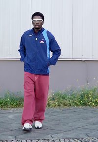 Blå og lyseblå zip-up jakke med logo, lyserøde brede bukser og hvide sneakers. Reflekterende solbriller fuldender outfittet.
