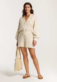 Abito corto beige chiaro con motivo floreale testurizzato, vita elastica e maniche lunghe. Accessoriato con una borsa a rete intrecciata e sandali flat.