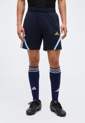 Atleta maschile che indossa pantaloncini da calcio blu navy con accenti azzurri, calzettoni blu navy coordinati con strisce bianche e scarpe da calcio nere.