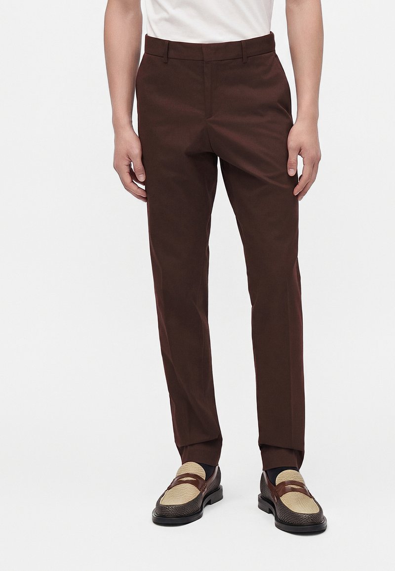 Homme portant un pantalon slim marron et des mocassins bicolores marron et beige, debout sur fond blanc.