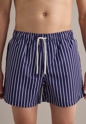 Torso masculino con pantalones cortos de baño azul marino con rayas blancas verticales y un cordón blanco atado en la cintura.