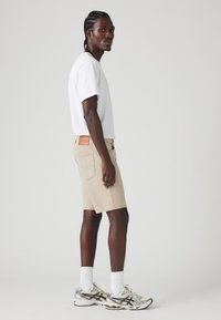 Shorts din denim bej cu o croială dreaptă, asortați cu un tricou simplu alb și adidași metalici pentru alergare. Detalii notabile de cusătură pe pantaloni.